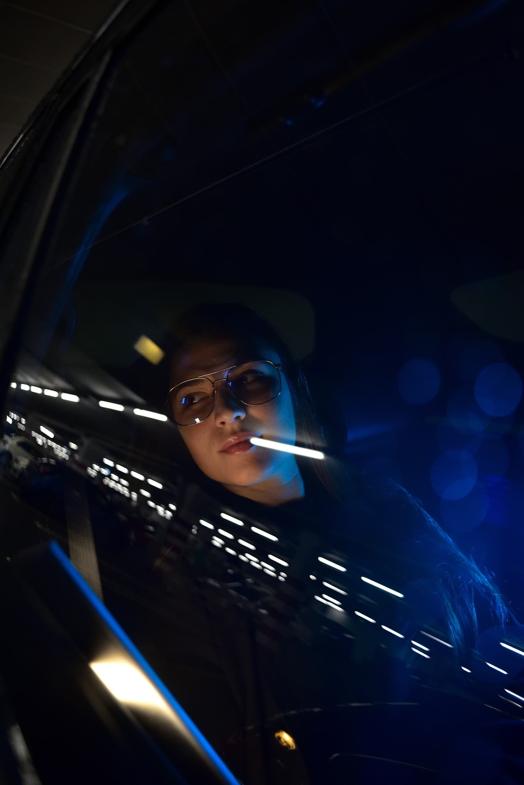 Frau mit Brille im Auto bei Nacht, umgeben von blauem Lichtbokeh